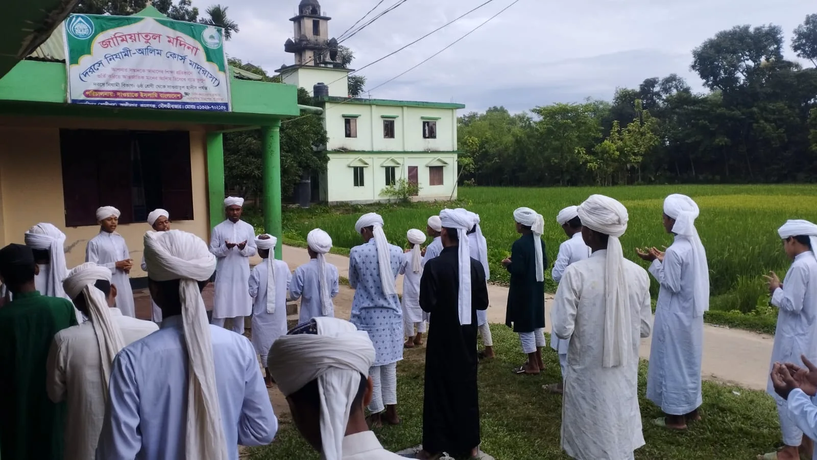 Students in white caps standing outdoors with their hands raised in prayer beside a building with green pillars