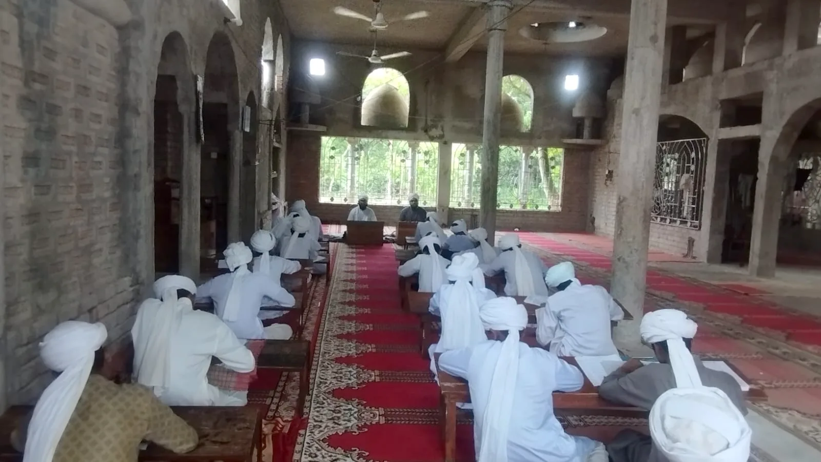 Students seated at low desks reading inside a mosque hall with a red carpet