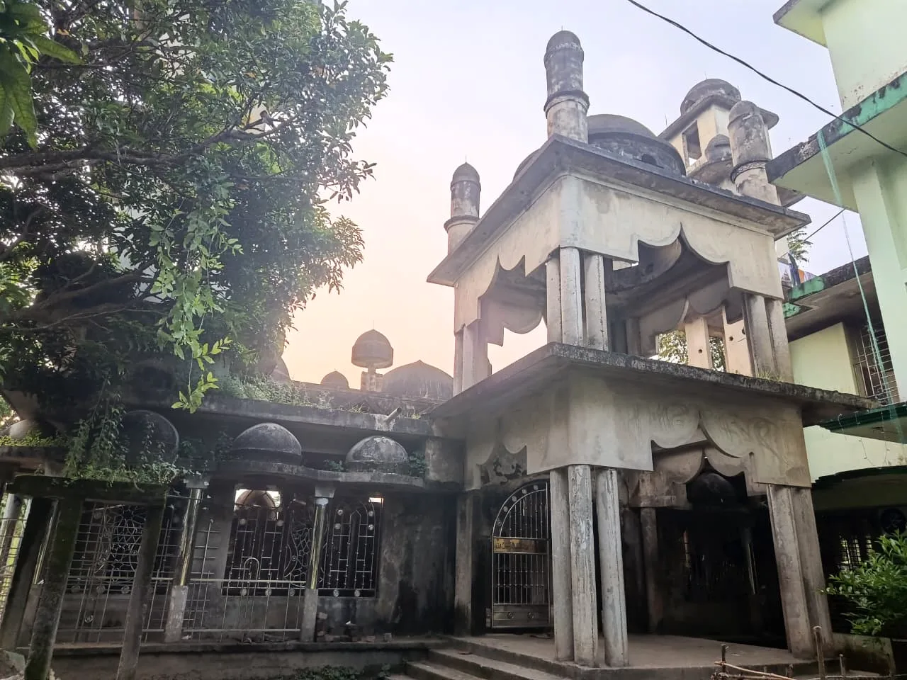 An old weathered mosque with small domes and a two-tier gateway, surrounded by trees