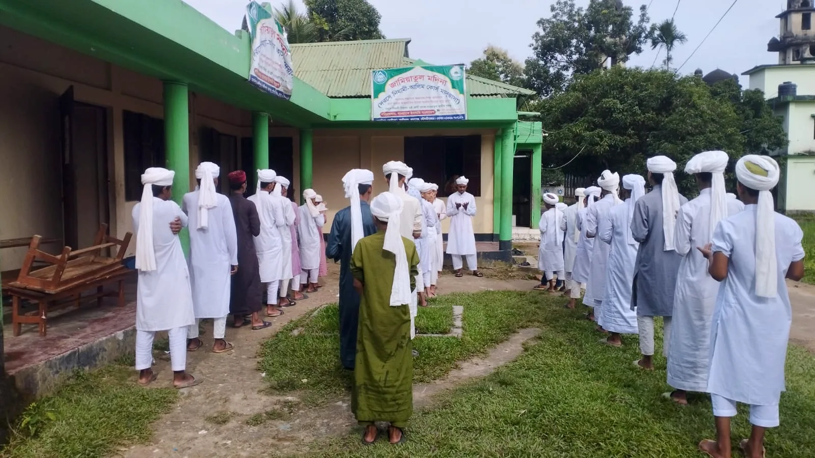 Students standing in rows outdoors in front of a single-storey building with green pillars and banners