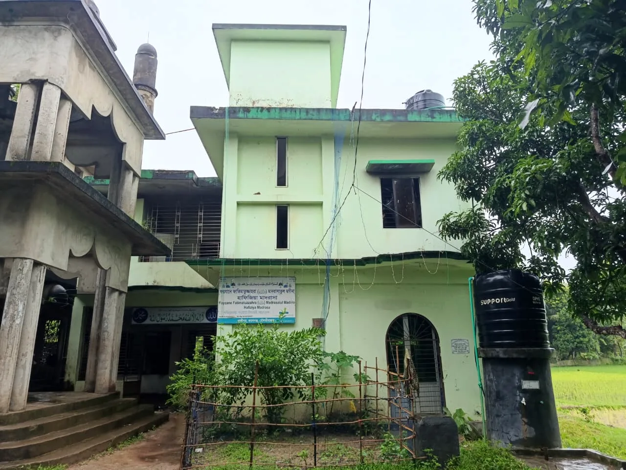 A multi-storey pale green building with a signboard, beside an old mosque