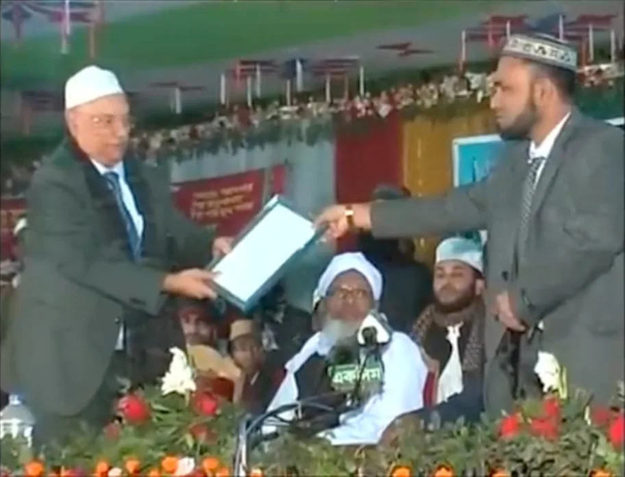 Television footage of a man in a suit handing a document to another man on a stage decorated with flowers