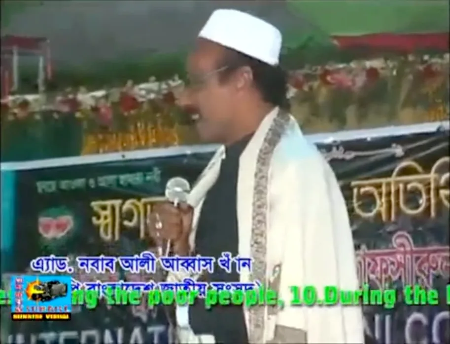 Television footage of a speaker in a white cap talking into a microphone, with a Bengali name caption